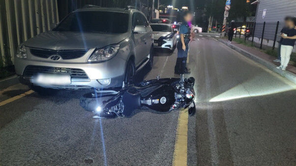 야간 도로에 은색 SUV 차량 앞에 검은 오토바이가 넘어져 있고, 몇 명의 사람들이 주변에 서 있는 모습