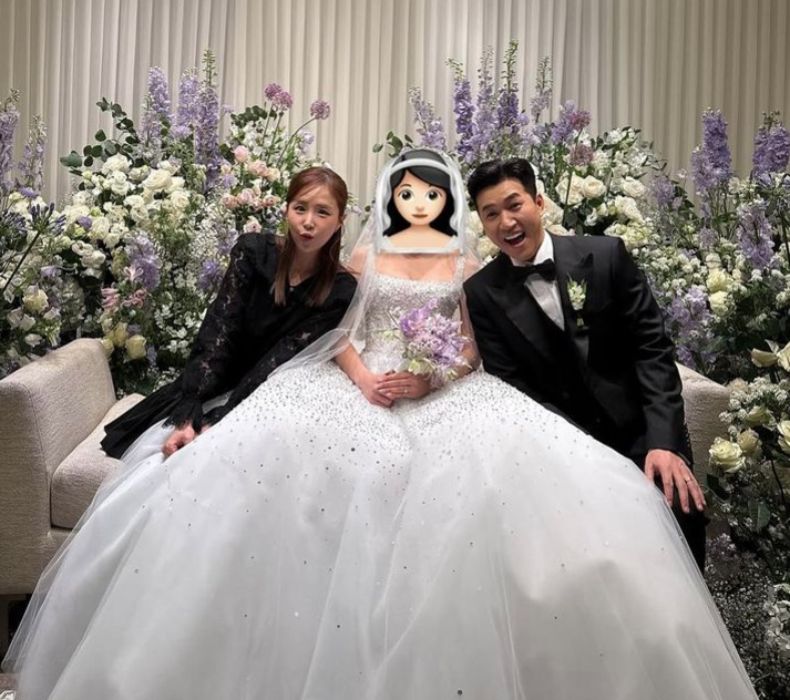 화려한 꽃 장식 앞에서 신부의 얼굴이 이모지로 가려진 채 웨딩드레스를 입은 사람과 턱시도, 검정 드레스를 입은 두 사람이 소파에 앉아 기념사진을 찍고 있다.