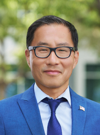 짧은 머리와 안경을 쓴 남성이 파란색 정장과 넥타이를 입고 미소를 지으며 서 있다. 배경에는 나무와 건물이 흐릿하게 보인다.