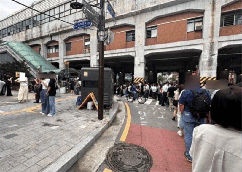 지하철역 근처에서 많은 사람들이 줄을 서 있는 모습. 일부 사람들은 자전거를 타고 지나가며, 주변에는 건물과 안내 표지판이 보임.