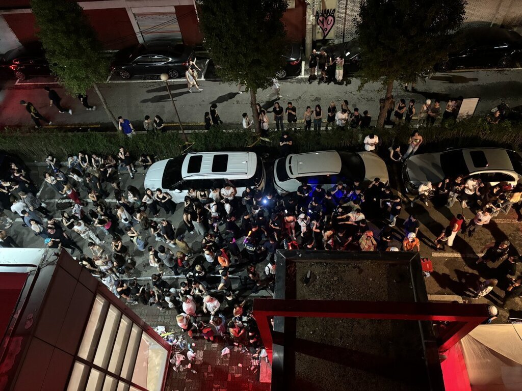 밤에 거리에서 많은 사람들이 모여 있는 모습이 위에서 내려다보이는 각도로 촬영됨. 길가에는 차량들이 주차되어 있고, 사람들은 무리를 지어 대화를 나누거나 이동하고 있음.