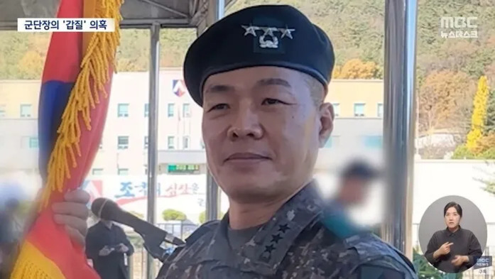 군복을 입은 남성이 군모를 쓰고 국기를 들고 있는 모습. 화면 좌측 상단에는 '군단장의 갑질 의혹'이라는 자막이 있고, 우측 하단에 수어 통역사가 있다.