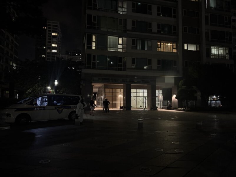 밤에 아파트 단지 입구 앞에 경찰차가 서 있고, 몇 명의 사람들이 건물 입구 근처에 모여 있다. 주변은 어둡고 일부 창문에는 불이 켜져 있다.
