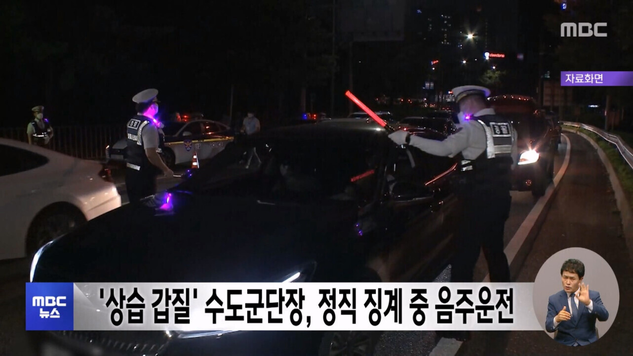 밤에 경찰관들이 도로에서 음주운전 단속을 하고 있으며, 한 경찰이 빨간 신호봉을 들고 차량 운전자와 대화하고 있다. 화면 하단에는 '상습 갑질' 수도군단장, 정직 징계 중 음주운전이라는 뉴스 자막과 수어 통역사가 보인다.