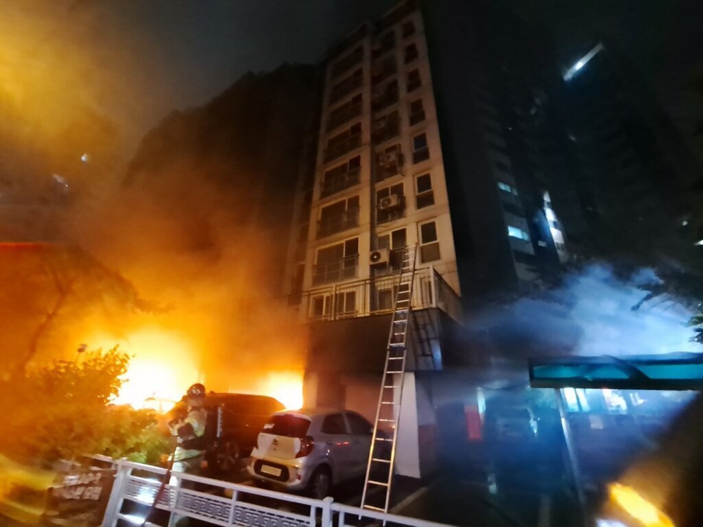 밤에 아파트 건물 1층에서 큰 불이 나 연기와 화염이 치솟고 있으며, 소방관이 진화 작업을 하고 있다. 사다리가 건물 외벽에 세워져 있다.