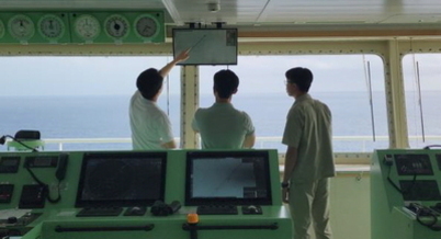 선박 조타실에서 세 명의 남성이 모니터를 보며 토론하고 있다. 창문 너머로 바다가 보인다.