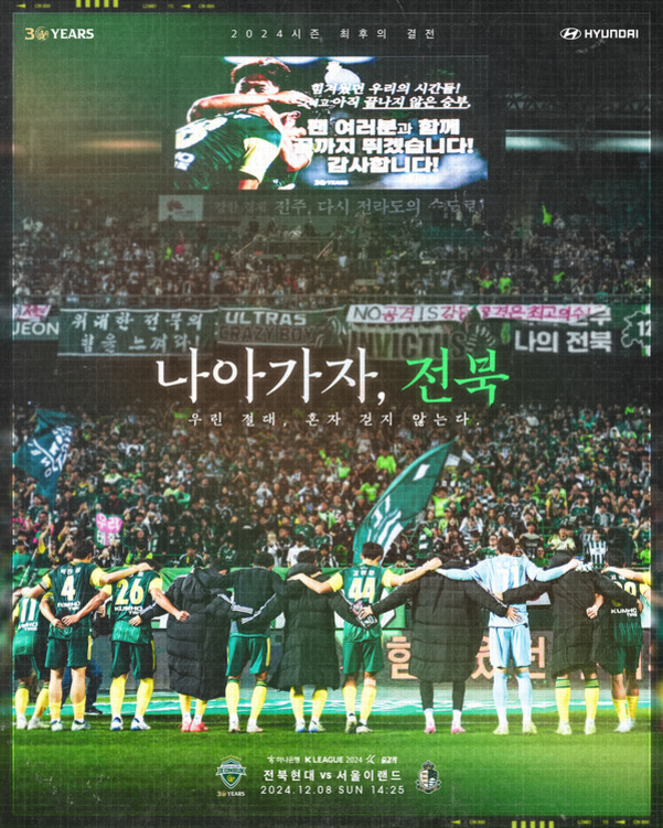 축구 경기장에서 전북현대 선수들이 팬들과 함께 팔짱을 끼고 서 있으며, 관중석에는 많은 팬과 응원 깃발이 보인다. 상단 전광판에는 메시지가 표시되어 있고, 중앙에 '나아가자, 전북'이라는 문구와 경기 정보가 적혀 있다.