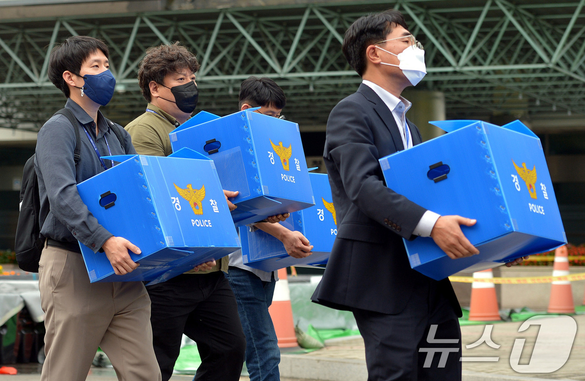여러 명의 남성이 경찰 로고가 있는 파란색 상자를 들고 이동하고 있다. 모두 마스크를 착용하고 있으며 공식적인 분위기이다.
