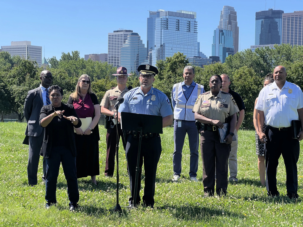 도시의 고층 빌딩이 배경인 공원에서 경찰, 경관 및 다양한 사람들이 모여 연설을 듣고 있고 한 사람이 수화로 통역하고 있는 모습.