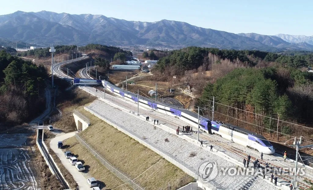 산과 숲이 배경인 시골 지역에서 고속열차가 탈선하여 선로 옆에 정차해 있고, 여러 명의 사람들이 현장에 모여 있다.