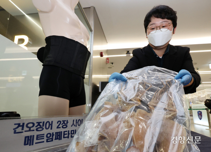 마스크와 장갑을 낀 사람이 비닐봉지에 든 건오징어를 들고 있고, 옆에는 허리에 벨트를 착용한 마네킹이 있다. 앞쪽 표지판에는 '건오징어 2장 사이 메트암페타민'이라는 문구가 보인다.