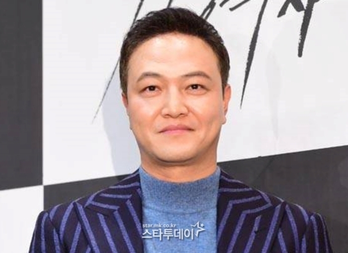 줄무늬 정장을 입고 미소짓는 남성이 행사 포토월 앞에 서 있다.