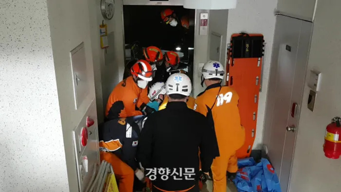 여러 명의 소방대원과 구급대원이 복도에서 구조 작업을 하고 있다. 한 대원이 들것에 환자를 싣고 있으며, 벽에는 긴급 장비와 의료 가방이 놓여 있다.