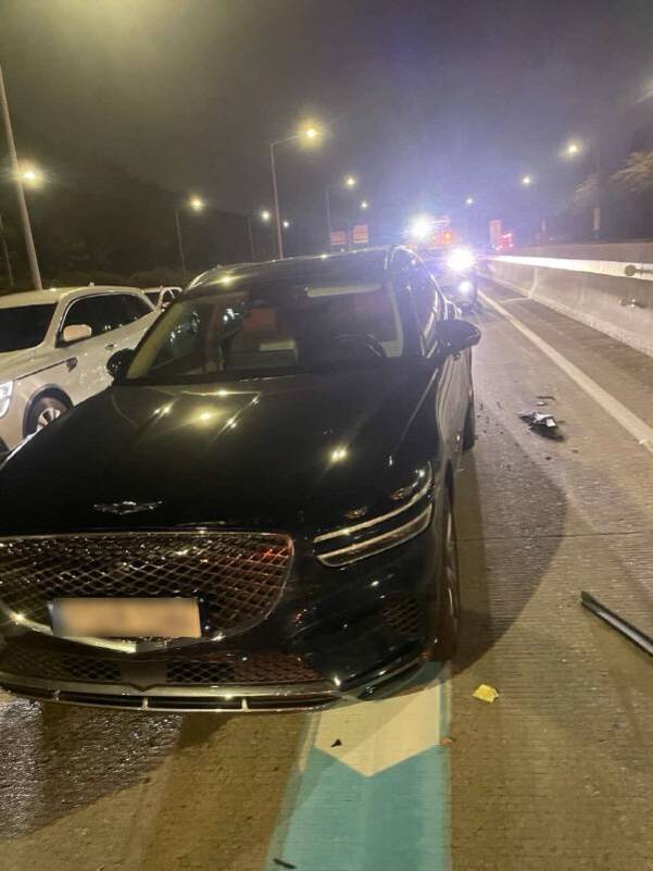 야간 고속도로에서 검은색 차량이 앞부분에 손상을 입고 정차해 있으며, 주변에는 다른 차량과 파손된 자동차 부품들이 보인다.