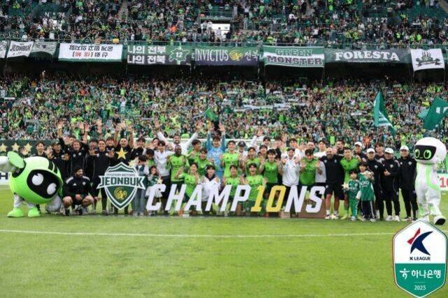 축구 경기장에서 전북 현대 모터스 선수단과 마스코트들이 'CHAMPIONS'라는 대형 글자를 들고 우승을 기념하며 단체 사진을 찍는 모습. 관중석에는 많은 팬들이 초록색 옷을 입고 응원하고 있다.