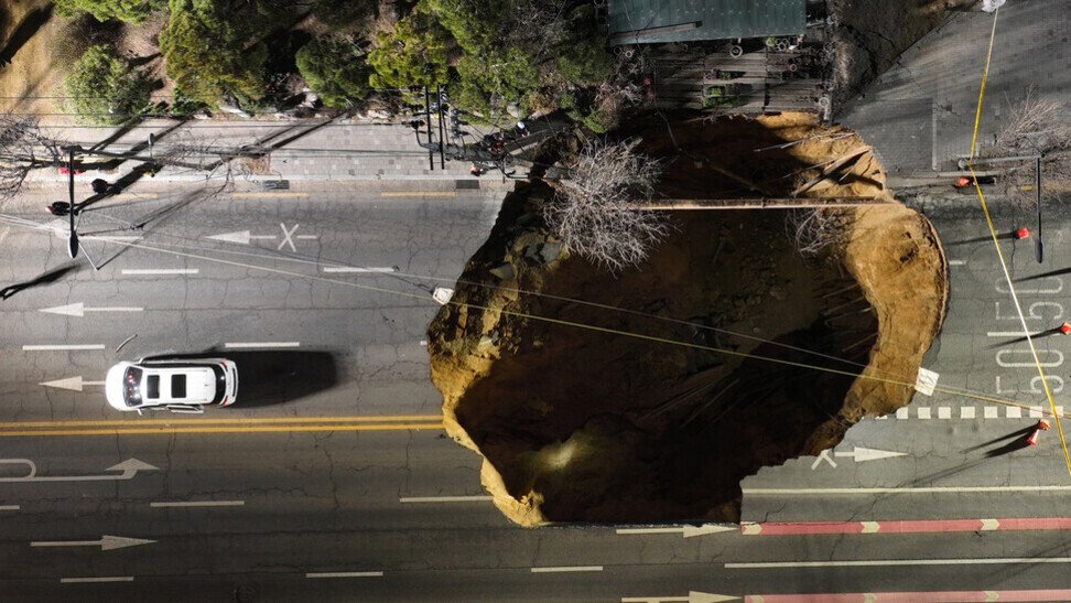 도로 한가운데에 대형 싱크홀이 생긴 모습과 그 옆을 지나가는 흰색 차량이 보인다.