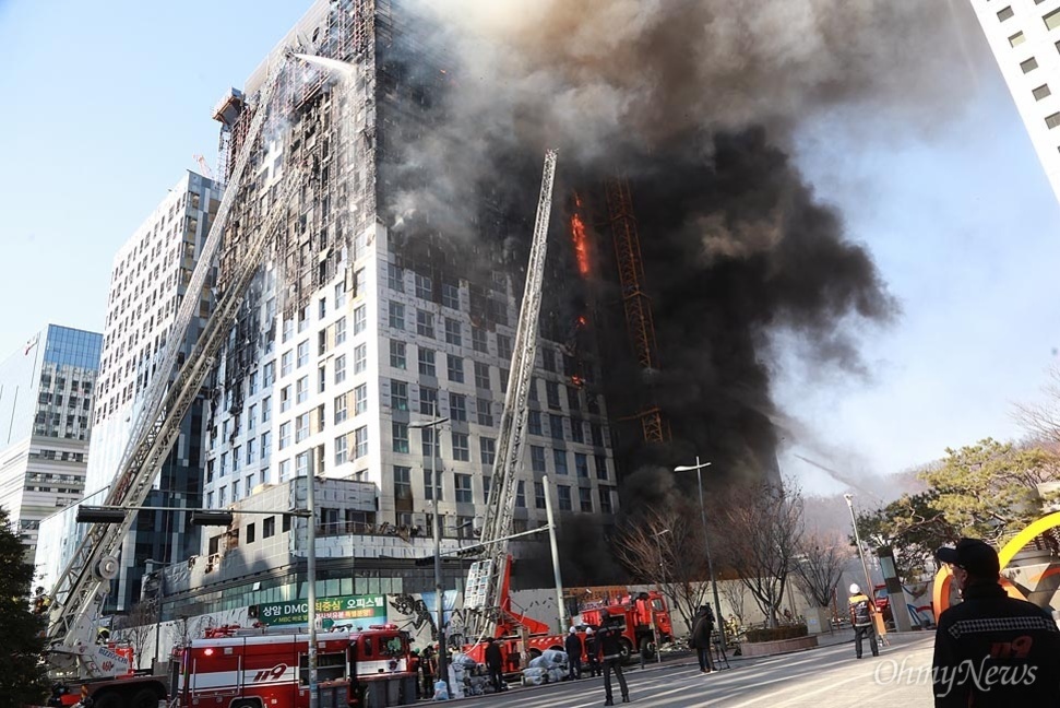 고층 건물에 큰 화재가 발생해 검은 연기가 치솟고 있으며, 소방차와 소방관들이 출동해 진화 작업을 하고 있는 모습