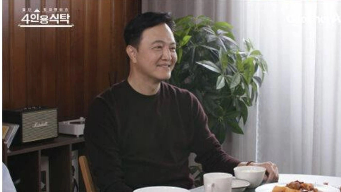 갈색 상의를 입은 남성이 식탁에 앉아 웃으며 대화를 나누고 있다. 테이블 위에는 음식과 그릇이 놓여 있고, 배경에는 화분과 커튼이 보인다.