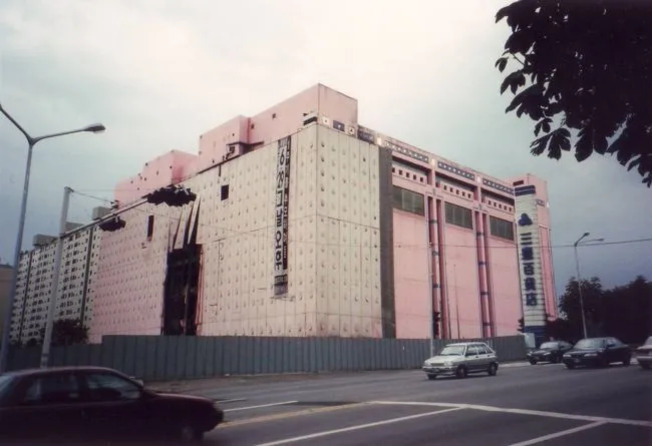 분홍색 외벽을 가진 대형 건물이 도로 옆에 위치해 있고, 몇 대의 자동차가 지나가는 모습