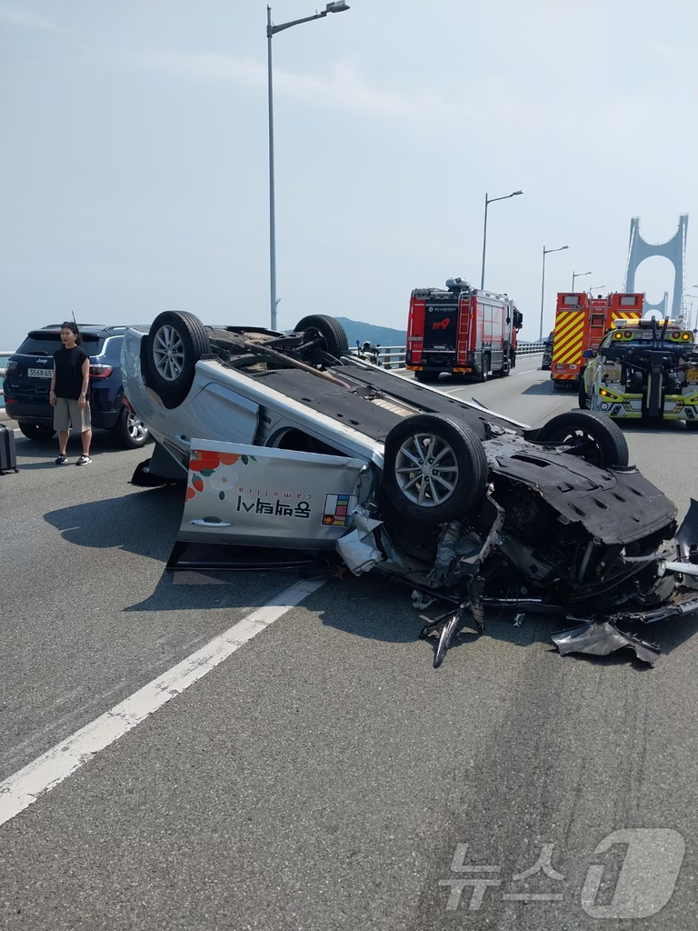 도로 위에 차량 한 대가 전복되어 뒤집혀 있고, 주변에는 소방차와 구조대 차량이 정차해 있다. 한 사람이 사고 현장 근처에 서 있으며, 멀리 다리가 보인다.