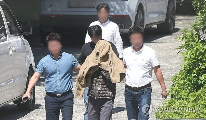 남성 한 명이 겉옷으로 얼굴을 가린 채 두 남성에게 양쪽 팔을 붙잡혀 이동하고 있으며, 주변에는 차량과 다른 사람들이 있다.