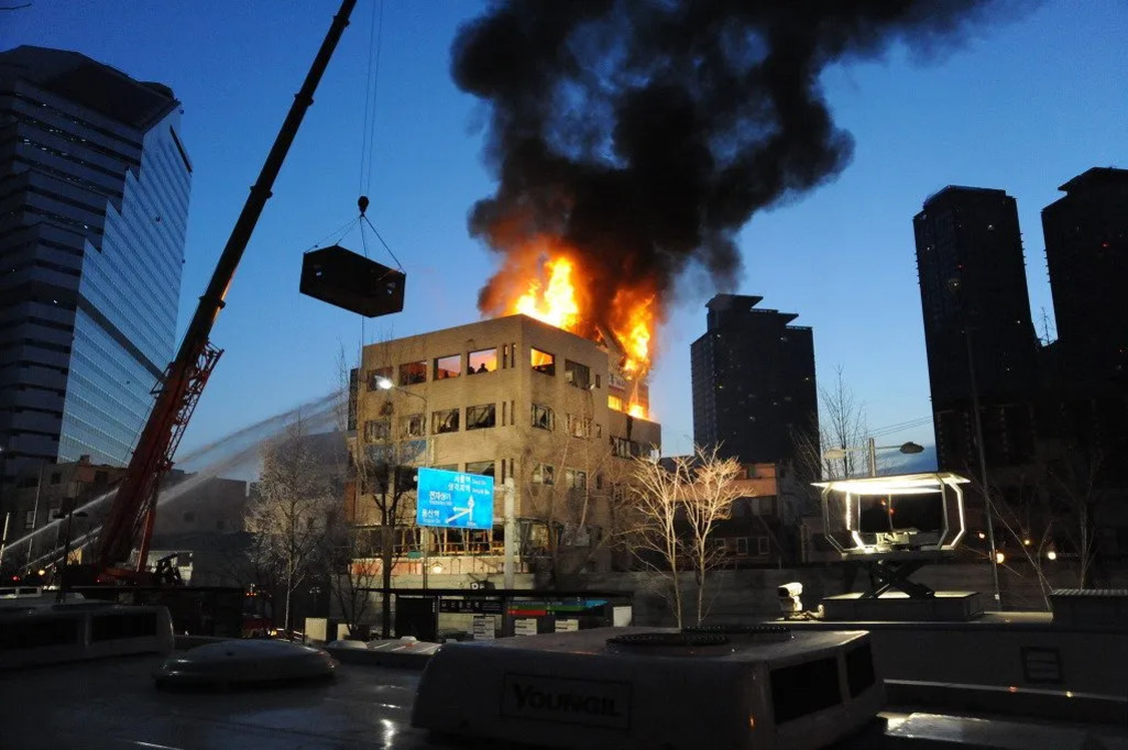 도심의 저녁 시간, 4층 건물에서 큰 불길과 검은 연기가 치솟고 있으며 소방차가 물을 뿌려 화재를 진압하고 있다. 크레인이 구조 작업 중이다.