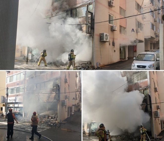 소방관들이 연기가 자욱하게 피어오르는 건물 화재 현장에서 진압 작업을 하고 있다. 건물 외벽이 검게 그을렸고, 주변에 잔해가 널려 있다.