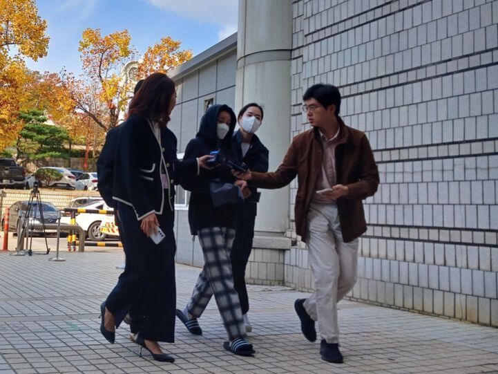 가을 풍경 속에서 네 명의 사람들이 건물 입구로 걸어가는 모습. 한 사람은 후드와 마스크를 착용했고, 다른 두 명이 그 옆을 에워싸고 있으며 한 남성이 인터뷰하듯 손에 무언가를 들고 있다.