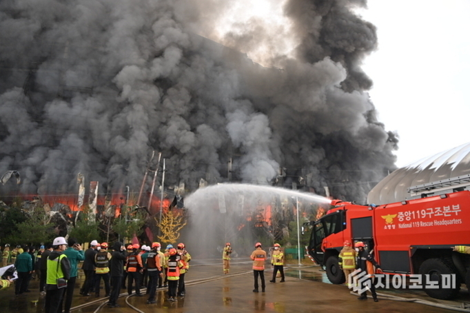 대형 화재 현장에서 소방관들이 진화 작업을 하고 있으며, 검은 연기가 하늘로 치솟고 있다. 빨간색 중앙119구조본부 소방차가 물을 뿌리고 있고 여러 명의 구조대원이 주변에 모여있다.