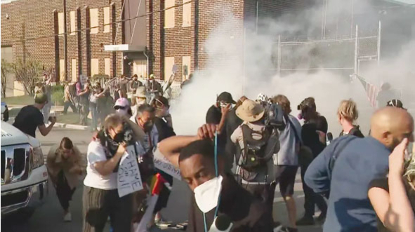 여러 명의 시위대가 건물 앞에서 최루탄 연기 속에 고개를 숙이고 도망치거나 얼굴을 가리고 있다.