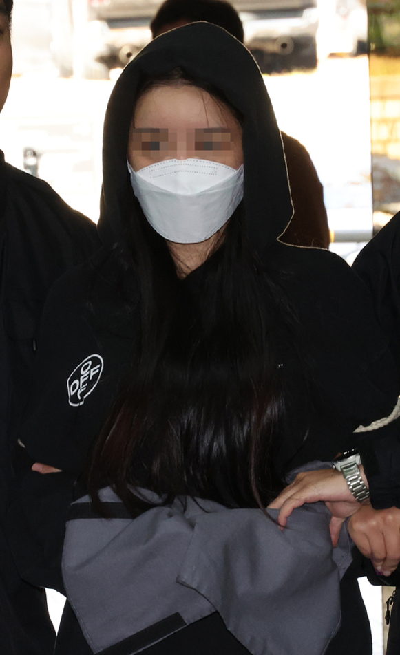 후드티와 마스크를 착용하고 얼굴이 모자이크 처리된 여성이 양옆 사람에게 팔짱을 낀 채 이동하는 모습