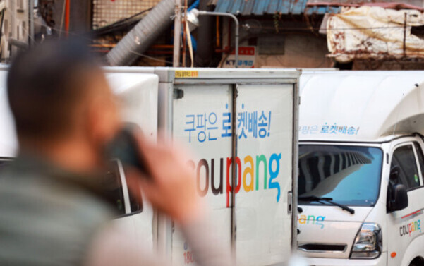 쿠팡 로켓배송 트럭 두 대가 도로에 주차되어 있고, 전면에는 흐릿하게 남성이 휴대전화를 들고 있다.