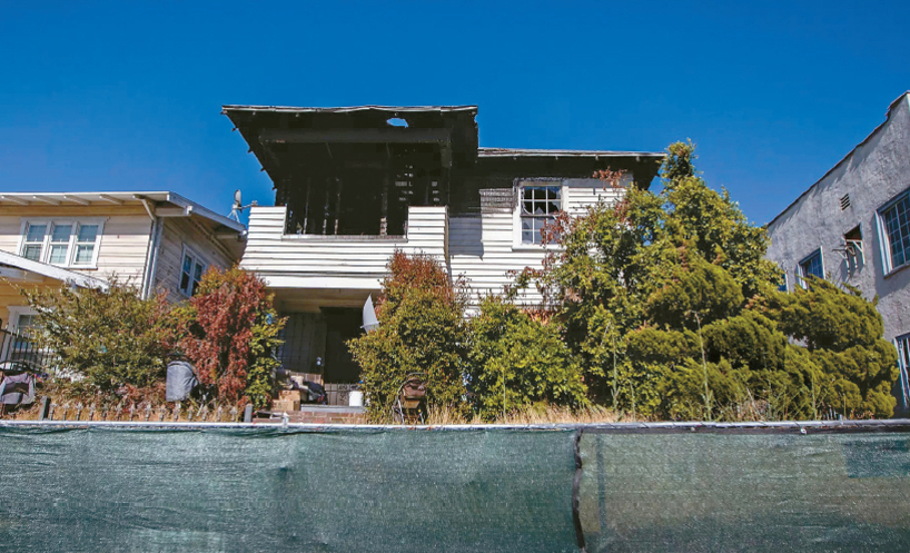 불에 탄 흔적이 남아 있는 2층 주택과 그 앞의 무성한 덤불, 파란 하늘