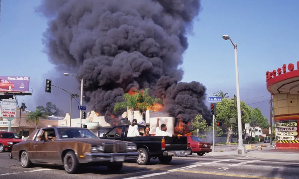 도심 교차로에서 큰 화재가 발생해 검은 연기가 하늘로 치솟고 있으며, 차량과 사람들이 그 앞을 지나가는 모습