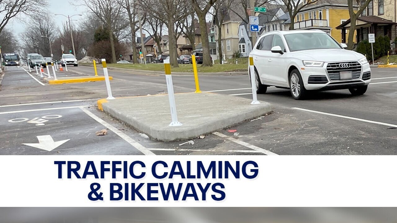 교통 진정 구역과 자전거 도로가 설치된 도시 거리, 흰색 SUV 차량이 지나고 있고 도로에는 분리대와 안내 표지판이 보임.