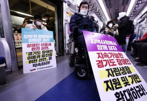 지하철 객차 안에서 휠체어를 탄 장애인과 다른 시민이 장애인 이동권 보장과 지원 확대를 요구하는 피켓을 들고 시위를 하고 있다. 모두 마스크를 착용하고 있으며, 지하철 문 근처와 통로에 위치해 있다.