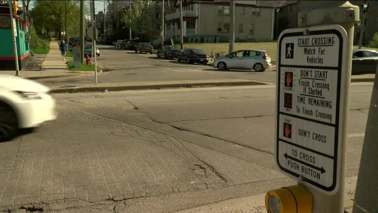 횡단보도 버튼과 안내 표지판이 있는 교차로, 도로를 지나가는 차량과 인도가 보인다.