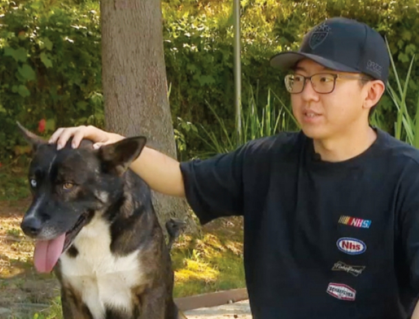 안경을 쓴 남성이 검은색과 흰색이 섞인 큰 개의 머리를 쓰다듬고 있다. 배경에는 나무와 풀숲이 보인다.