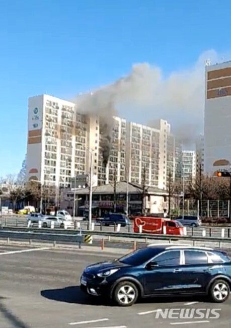 고층 아파트 건물에서 검은 연기가 크게 피어오르고 있으며, 도로에는 차량들이 지나가고 있다.