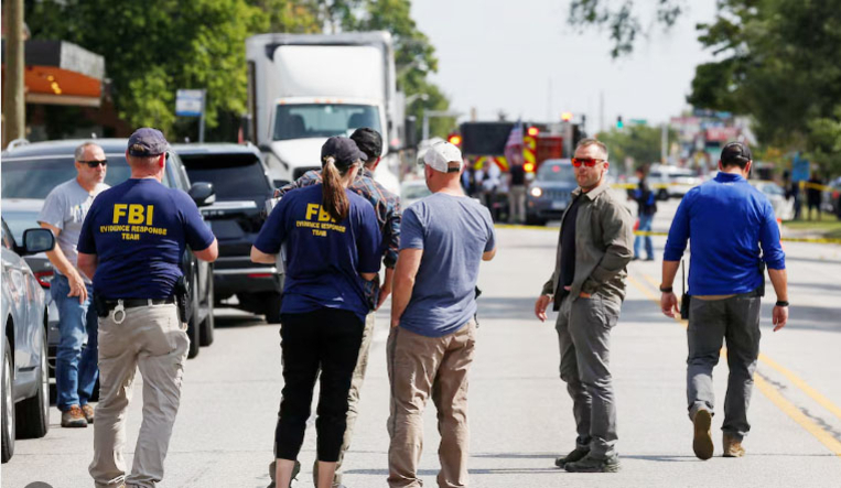 도로 위에 FBI 요원들과 여러 명의 사람들이 모여 있으며, 뒤쪽에는 경찰차와 구급차가 보인다.
