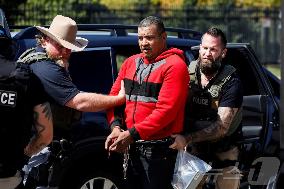 경찰관 두 명이 수갑을 찬 남성을 호송하고 있다. 한 경찰은 모자를 썼으며, 차량 앞에서 상황이 진행되고 있다.