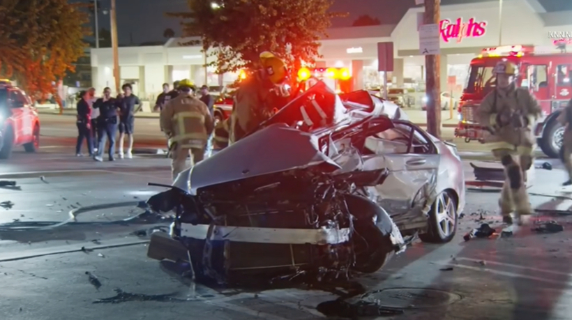 심하게 파손된 자동차와 이를 둘러싼 소방관들, 배경에는 구급차와 사람들이 보임.