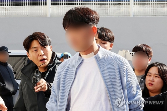 여러 명의 사람들이 야외에서 모여 있으며, 앞쪽 남성의 얼굴이 흐릿하게 처리되어 있다. 한 사람이 마이크를 들고 인터뷰 중인 모습이다.