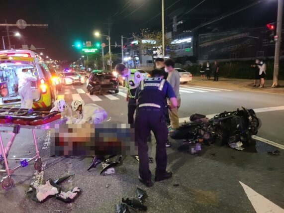 야간 도로에서 오토바이 사고 현장. 경찰과 구급대원이 부상자를 구조하고 있으며, 주변에 파손된 오토바이와 차량, 구급차가 보인다.